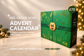 A green Christmas Advent Calendar box with gold festive illustrations, displayed on a marble surface with holiday lights and pine branches. 